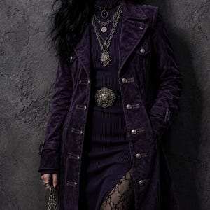 May include: A long, dark purple velvet coat with silver buttons and a matching belt. The coat is worn over a dark purple dress and layered necklaces. The look is completed with black lace stockings and a black handbag.