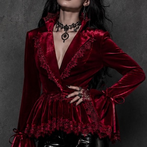 May include: A deep red velvet jacket with intricate lace trim and bell sleeves. The jacket features a V-neckline and is paired with black leather pants. A black choker necklace and rings complete the gothic-inspired ensemble.