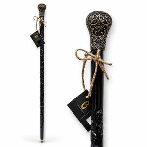 May include: A black walking stick featuring a metallic, dark silver-toned handle with an intricate floral relief pattern. The shaft is carved with a continuous vine design and is tied with a brown twine string holding a black rectangular tag.