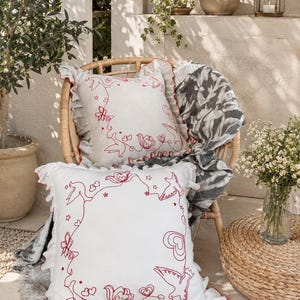 May include: Two square throw pillows with ruffled edges and red embroidered line art featuring birds, flowers, hearts, and stars. The pillows are made of white fabric, and the embroidery creates a decorative frame around the center of each.