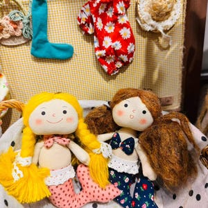 May include: Two plush mermaid dolls with embroidered faces and yarn hair sit in a polka dot basket. One doll has yellow hair and a pink patterned tail, while the other has brown hair and a dark blue floral tail. Miniature doll clothes hang above.