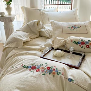 May include: A cream-colored bedding set featuring delicate floral embroidery in shades of red, blue, and green. The set includes a duvet cover, pillowcases with the text Hand Embroidered, and a cylindrical bolster pillow, all arranged on a bed.