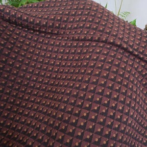 May include: A dark brown textile featuring a repeating geometric pattern of small black triangles and tan dots arranged in a grid. The fabric displays a textured surface with a visible seam and a section of decorative stitching along the edge.