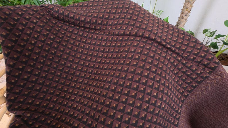 May include: A dark brown textile featuring a repeating geometric pattern of small black triangles and tan dots arranged in a grid. The fabric displays a textured surface with a visible seam and a section of decorative stitching along the edge.