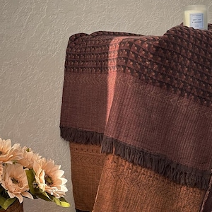 May include: A rectangular throw blanket featuring a gradient of deep burgundy to burnt orange tones. The fabric displays a textured pattern of small geometric shapes and chevron lines, finished with dark brown fringe along the bottom edges.
