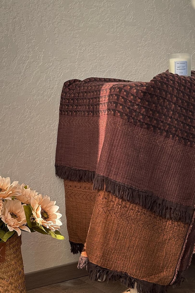 May include: A rectangular throw blanket featuring a gradient of deep burgundy to burnt orange tones. The fabric displays a textured pattern of small geometric shapes and chevron lines, finished with dark brown fringe along the bottom edges.