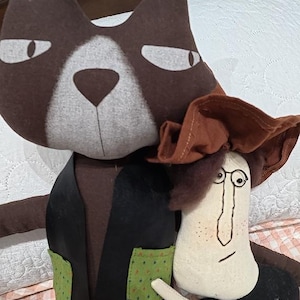 May include: A plush brown cat doll with a white face patch, wearing a black vest with two green pockets, standing next to a smaller cream-colored doll with a brown hat, round wire-rimmed glasses, and a black outfit, both made of fabric.