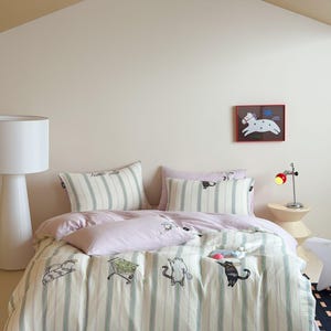 May include: A bedding set featuring a light cream and green striped pattern with embroidered cat illustrations. The set includes a duvet cover, two rectangular pillows, and one smaller pillow, all accented with playful cat motifs and text.
