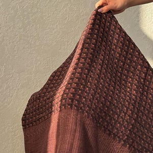 May include: A square textile featuring a dark brown and black geometric pattern of small squares with central dots. The fabric has a wide, solid dark brown border with a subtle chevron texture and a fringe edge along the bottom hemline.