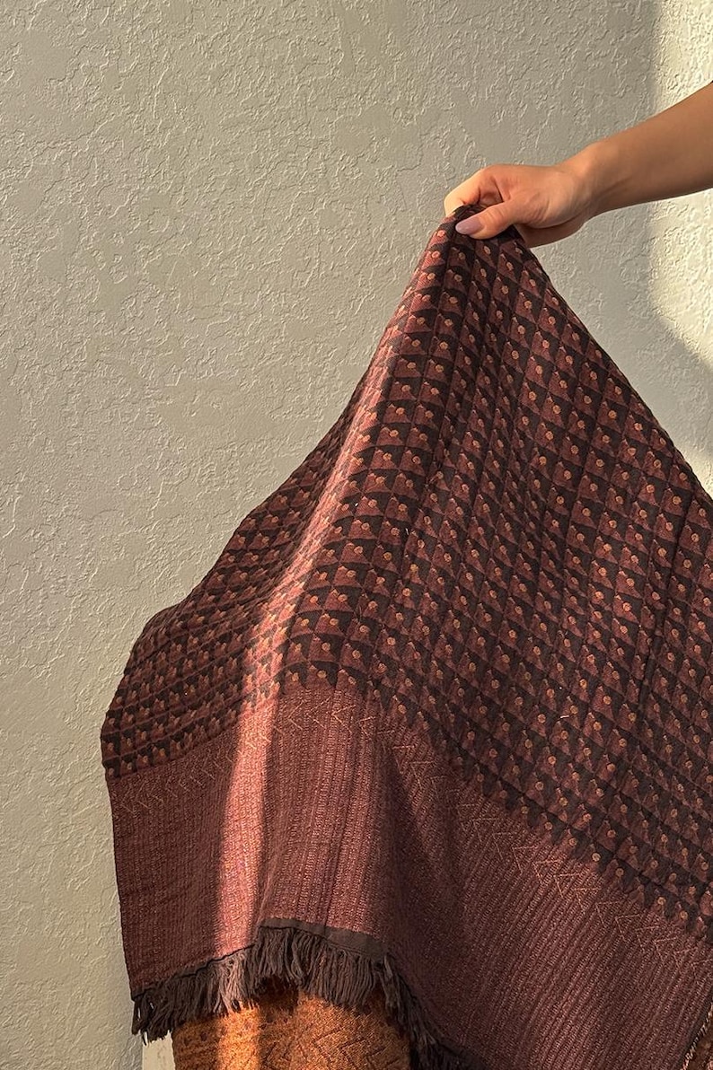 May include: A square textile featuring a dark brown and black geometric pattern of small squares with central dots. The fabric has a wide, solid dark brown border with a subtle chevron texture and a fringe edge along the bottom hemline.