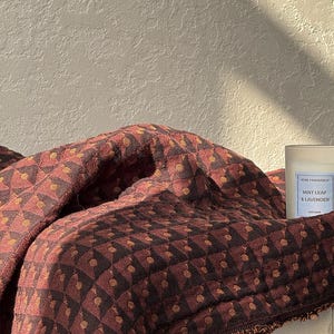 May include: A folded textile featuring a dark brown and rust-colored geometric pattern with small circular accents. Beside the fabric sits a clear glass candle jar with a white label reading HOME FRAGRANCE MINT LEAF & LAVENDER AROMA.