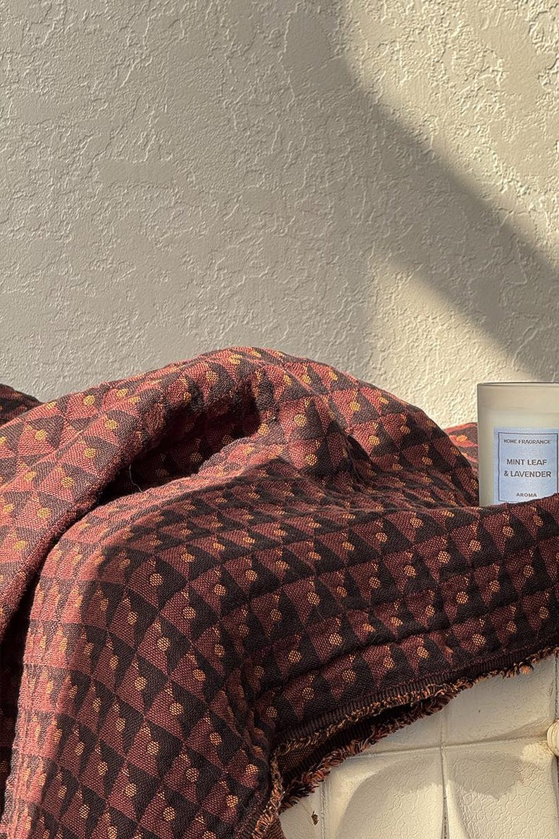 May include: A folded textile featuring a dark brown and rust-colored geometric pattern with small circular accents. Beside the fabric sits a clear glass candle jar with a white label reading HOME FRAGRANCE MINT LEAF & LAVENDER AROMA.
