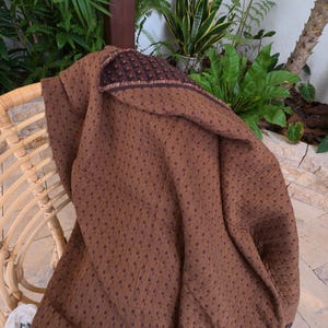 May include: A brown textile featuring a repeating pattern of small, dark brown dots. The fabric is draped over a curved, light-colored woven chair. The material has a textured appearance with a visible hemmed edge showing a darker brown border.