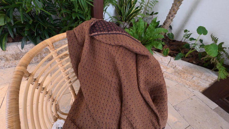 May include: A brown textile featuring a repeating pattern of small, dark brown dots. The fabric is draped over a curved, light-colored woven chair. The material has a textured appearance with a visible hemmed edge showing a darker brown border.