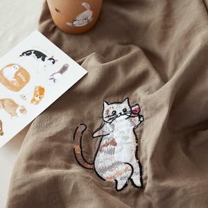 May include: A tan fabric cloth featuring an embroidered calico cat holding a small red object. Next to the cloth is a tan cup with a cat illustration and a white card displaying various cat drawings, including one with the text meow.