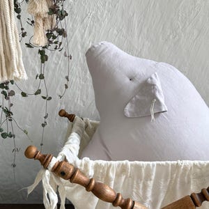 May include: A light gray, pig-shaped stuffed pillow with a small triangular ear and a tiny embroidered eye. The pillow rests inside a wooden cradle featuring turned posts and a white, textured fabric liner with delicate floral embroidery.