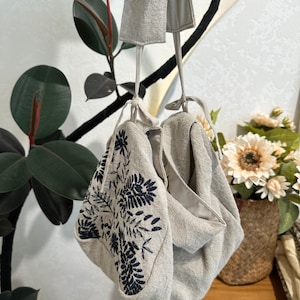 May include: A beige linen hobo bag featuring dark navy blue floral embroidery on the front panel. The bag has a soft, unstructured silhouette with a single top handle and tie closures at the top, showcasing a natural fabric texture throughout.