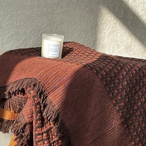 May include: A textured brown and orange throw blanket with fringe edges and a geometric pattern, featuring a small frosted glass candle with a white label reading Mint Leaf & Lavender placed on top of the folded fabric surface.