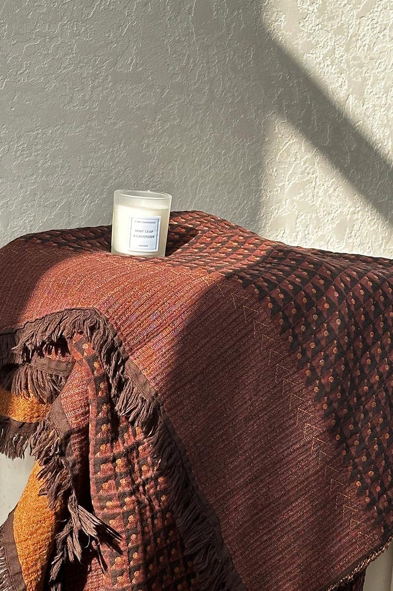 May include: A textured brown and orange throw blanket with fringe edges and a geometric pattern, featuring a small frosted glass candle with a white label reading Mint Leaf & Lavender placed on top of the folded fabric surface.