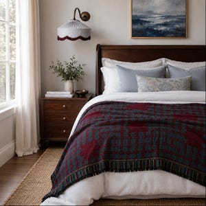 May include: A woven throw blanket featuring a dark red and teal grid pattern with a fringed edge, draped over a bed with white linens, light blue pillows, and a decorative lumbar pillow. A wooden nightstand sits beside the bed with a lamp.