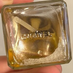 May include: A clear, square perfume bottle with the word "CHANEL" embossed on the bottom. The bottle contains a golden-coloured liquid and a white applicator. The bottle is held by a hand.
