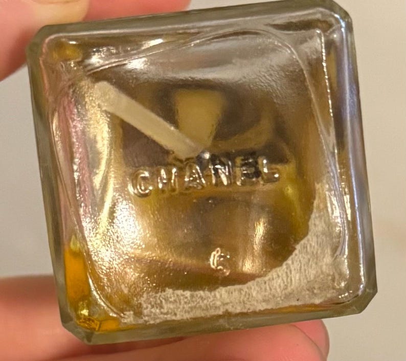May include: A clear, square perfume bottle with the word "CHANEL" embossed on the bottom. The bottle contains a golden-coloured liquid and a white applicator. The bottle is held by a hand.