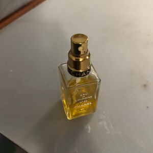 May include: A square glass perfume bottle with a gold-coloured cap. The bottle contains a light yellow liquid and has the words "EAU DE COLOGNE N&deg;22 CHANEL" printed on the front. The bottle is on a grey surface.