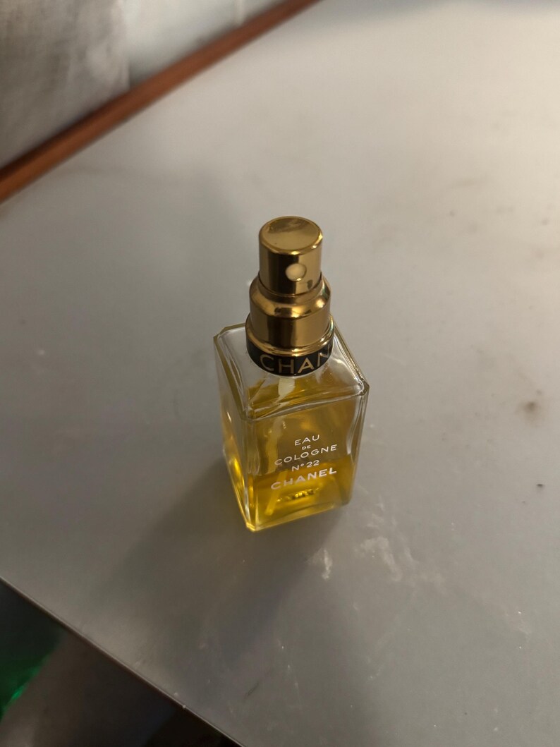 May include: A square glass perfume bottle with a gold-coloured cap. The bottle contains a light yellow liquid and has the words "EAU DE COLOGNE N&deg;22 CHANEL" printed on the front. The bottle is on a grey surface.