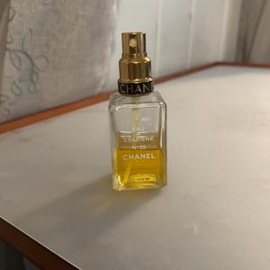 May include: A rectangular Chanel perfume bottle with a gold-coloured sprayer. The bottle contains a yellow liquid and has the words "EAU DE COLOGNE N&deg;22 CHANEL" printed on the front. The bottle is on a light-coloured surface.