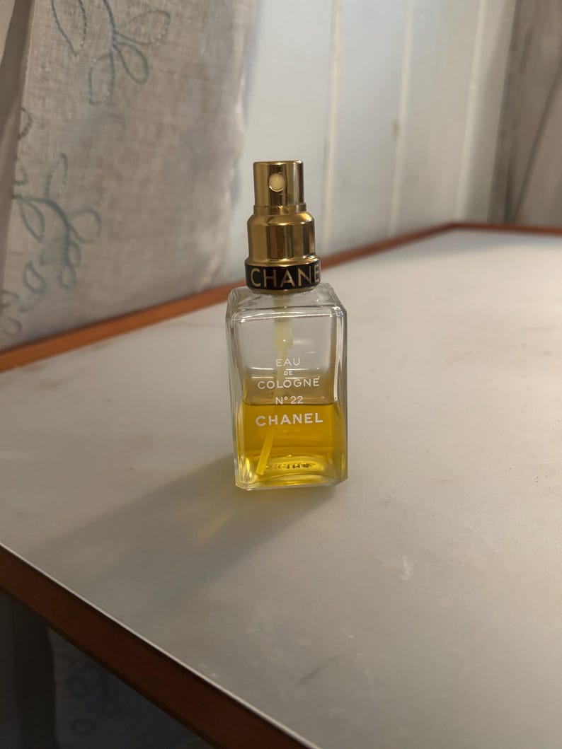 May include: A rectangular Chanel perfume bottle with a gold-coloured sprayer. The bottle contains a yellow liquid and has the words "EAU DE COLOGNE N&deg;22 CHANEL" printed on the front. The bottle is on a light-coloured surface.