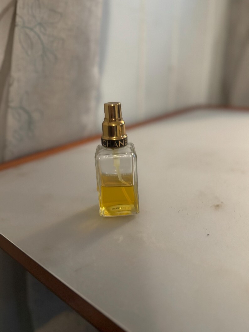 May include: A small, rectangular perfume bottle with a gold-coloured cap and the brand name "CHANEL" printed on it. The bottle contains a yellow liquid. The bottle is on a white surface.