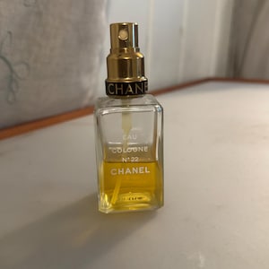 May include: A clear, rectangular perfume bottle with a gold-coloured spray top. The bottle contains a yellow liquid and features the text "EAU COLOGNE N&deg;22 CHANEL". The bottle is displayed on a light-coloured surface.