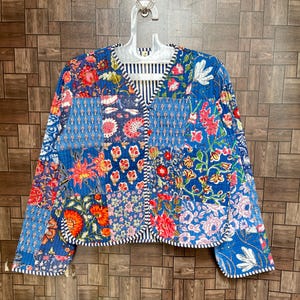 May include: A quilted jacket featuring a patchwork design of various blue, red, and white floral patterns. The garment has a V-neckline, long sleeves, and a front button closure, with a blue and white striped trim along the edges and collar.