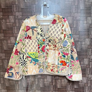 Op de afbeelding: Een doorgestikt jack met lange mouwen en een patchworkontwerp met diverse bloemen-, vogel- en geometrische patronen in tinten geel, rood, blauw en groen. Het jack heeft een V-hals, een opening aan de voorzijde en zichtbare stiksels in de stof.