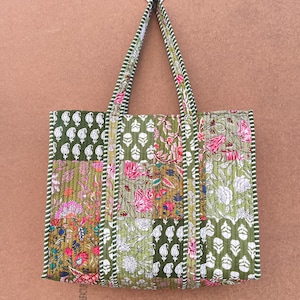 May include: A quilted tote bag featuring a patchwork design of various floral and paisley patterns in shades of olive green, pink, and white. The bag has two matching quilted handles with a green and white striped trim along the edges.