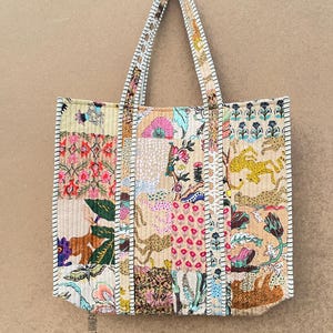 May include: A quilted tote bag featuring a patchwork design of various floral, animal, and geometric patterns in shades of pink, yellow, green, and tan. The bag has two long shoulder straps with a visible black and white whipstitch edge detail.