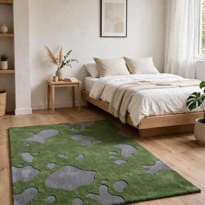 May include: A rectangular area rug featuring a deep green base color with an irregular, scattered pattern of gray, amoeba-like shapes. The rug has a textured, tufted surface and is positioned on a light-toned wooden floor in a bedroom setting.