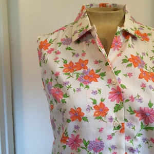 May include: A white sleeveless shirt with a collared neckline and a button-up front. The shirt has a floral print with pink, orange, and purple flowers.