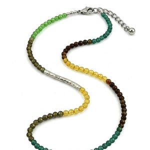 May include: A beaded necklace featuring a gradient of faceted green, brown, and yellow beads arranged in a curved pattern. The necklace includes a central curved silver-toned metal bar with a hammered texture and a silver-toned lobster claw clasp.