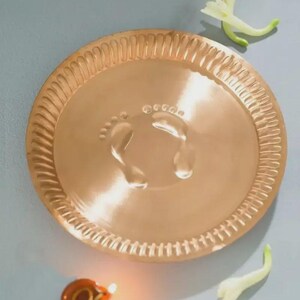 May include: A round, metallic copper-coloured plate featuring an embossed design of two small footprints in the centre and a decorative fluted edge. The plate is accompanied by a small, lit oil lamp and two white flower blossoms nearby.