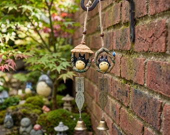May include: Two hanging wind chimes suspended from a black metal wall hook. Each features a small black and white creature figurine, a tan straw-like roof or branch frame, a metal leaf-shaped tag with Japanese text, and a small brass bell.