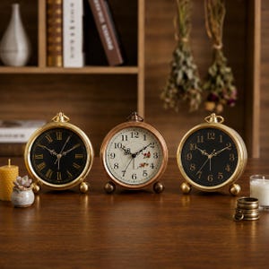 Könnte beinhalten: Drei runde Tischuhren mit Metallrahmen und Kugelfüßen. Zwei Uhren haben schwarze Zifferblätter mit goldenen Zahlen, eine mit römischen und eine mit arabischen Ziffern. Die mittlere Uhr hat ein weißes Zifferblatt mit arabischen Ziffern und Blumen.