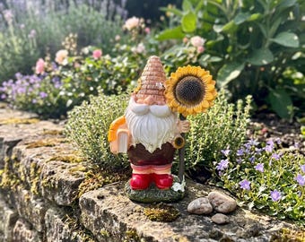 May include: A garden gnome figurine with a white beard, holding a sunflower and a book. The gnome wears a brown outfit, red boots, and a beehive-shaped hat. The figurine is set on a stone wall in a garden with flowers.