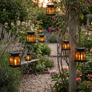 May include: A collection of black solar lanterns with amber glass panels are suspended from branches and a wooden post in a garden. Each lantern contains a lit candle, emitting a warm light. The garden includes flowers and a stone path.
