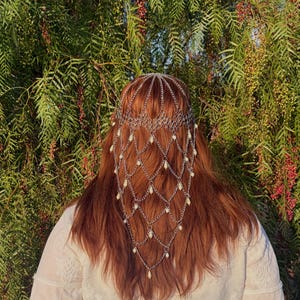 May include: A silver-toned metal headpiece featuring a delicate chain-link design that drapes over the hair. The accessory is adorned with small, teardrop-shaped white beads suspended at the intersections of the interconnected silver chains.