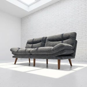 May include: A grey corduroy sofa with a two-cushion design. The sofa has a rounded shape and sits on four wooden legs. The background features a white brick wall and a white floor, with natural light coming from a skylight.