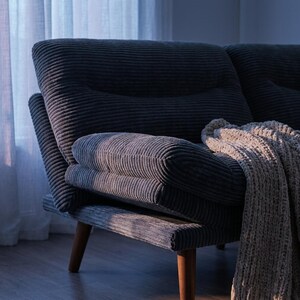 May include: A dark grey corduroy sofa with wooden legs. A beige knitted throw rests on the sofa. The sofa has a high back and armrests. The room has a window with a sheer white curtain.