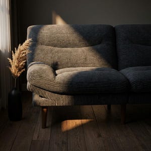 May include: A grey corduroy sofa with wooden legs. A black vase holds dried ornamental grass. The sofa is positioned on a wooden floor, with sunlight streaming in from a window, creating shadows across the scene.