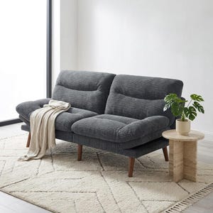 May include: A dark grey corduroy sofa with wooden legs. A beige throw is draped over the left side. A small round wooden side table holds a potted plant. The sofa sits on a cream-coloured rug.