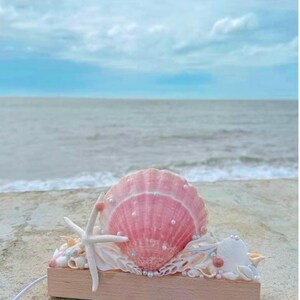 May include: A decorative light fixture featuring a large, central pink scallop shell adorned with small white pearls, flanked by a white starfish and various smaller seashells on a rectangular light-coloured wooden base with a white power cord.